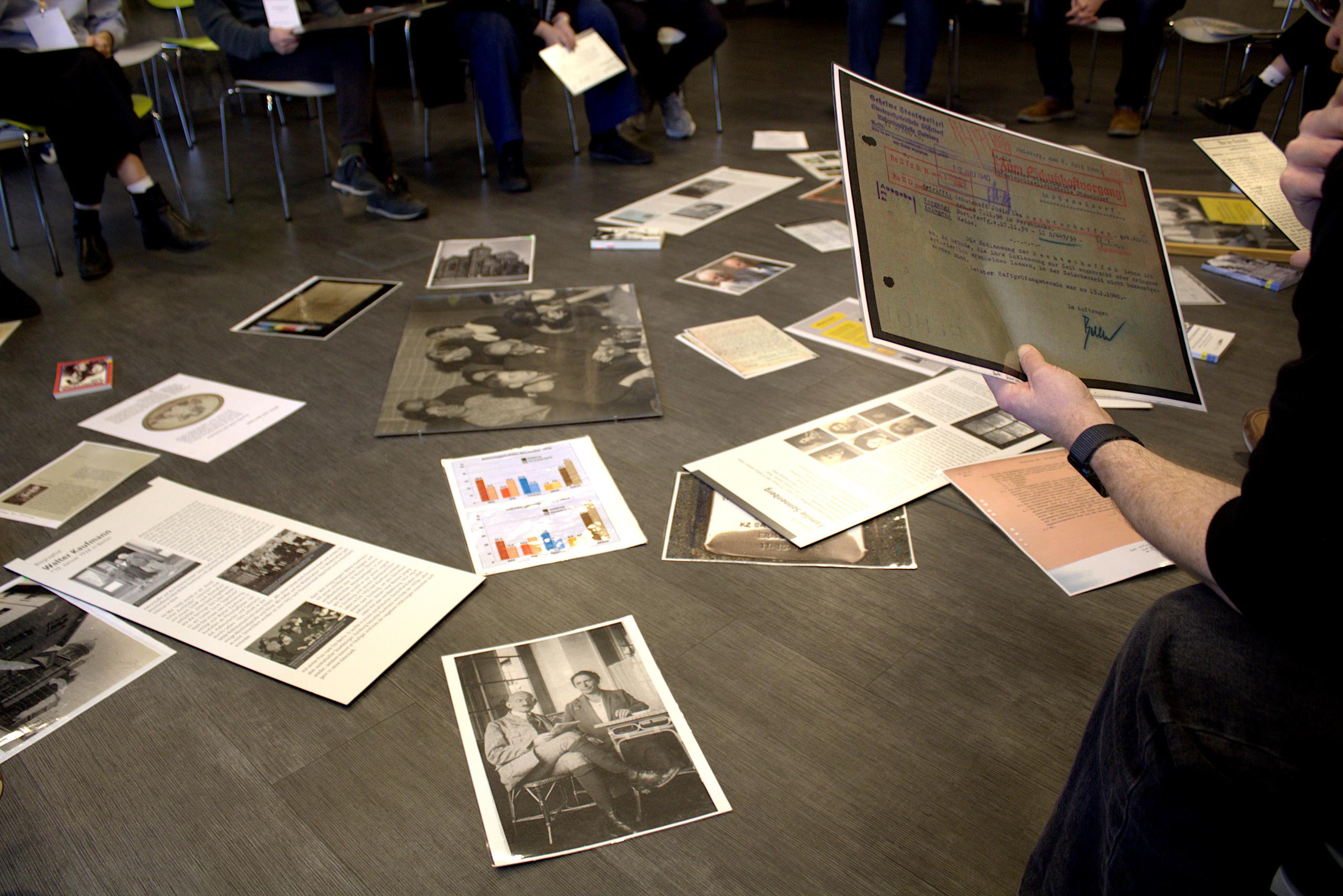 Auf dem Boden liegen verschiedene historische Quellen verstreut: Zeitungsartikel, Fotos und weitere biographische Quellen.
